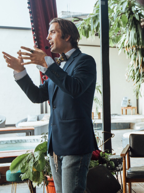 Shaman Peter in a navy suit and feathered bow tie, speaking beside a red velvet curtain